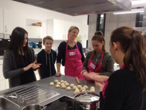 Mehrere Personen, darunter Jugendliche und Erwachsene mit pinkfarbenen Schürzen, stehen gemeinsam in einer modernen Küche und formen Teigbällchen auf einer Arbeitsfläche.